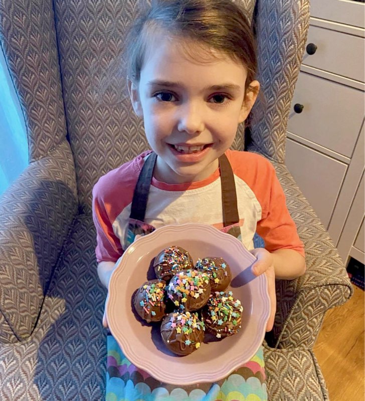 A little girl in an apron smiles and presents a bowl of chocolate and rainbow sprinkle baked treats.