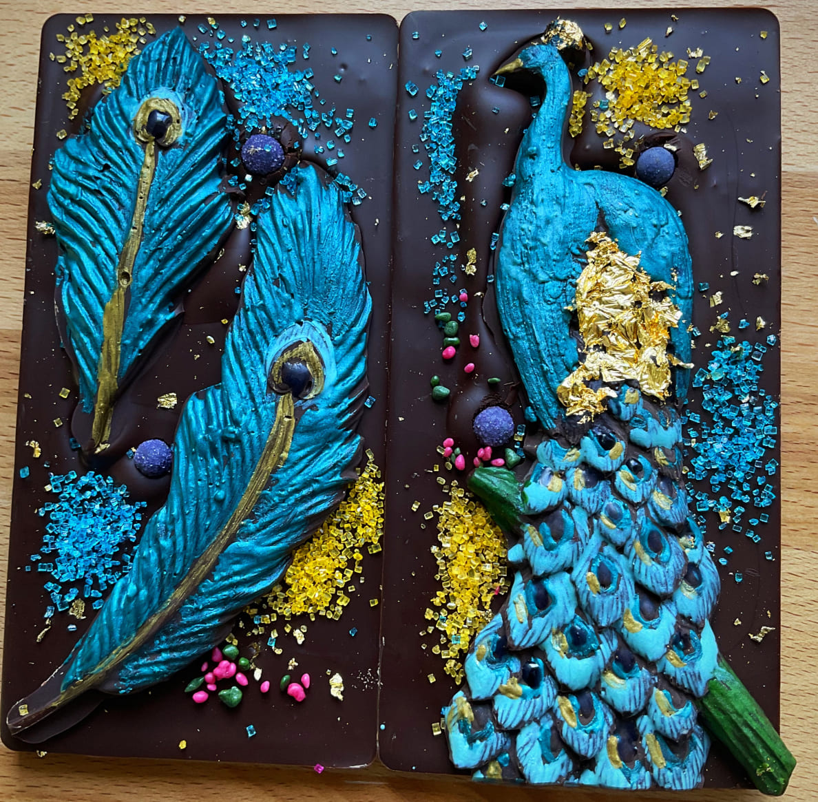A set of 2 chocolate bars from Raven Rising Global-Indigenous Chocolates are decorated with an edible blue and gold peacock design and a matching peacock feathers design.