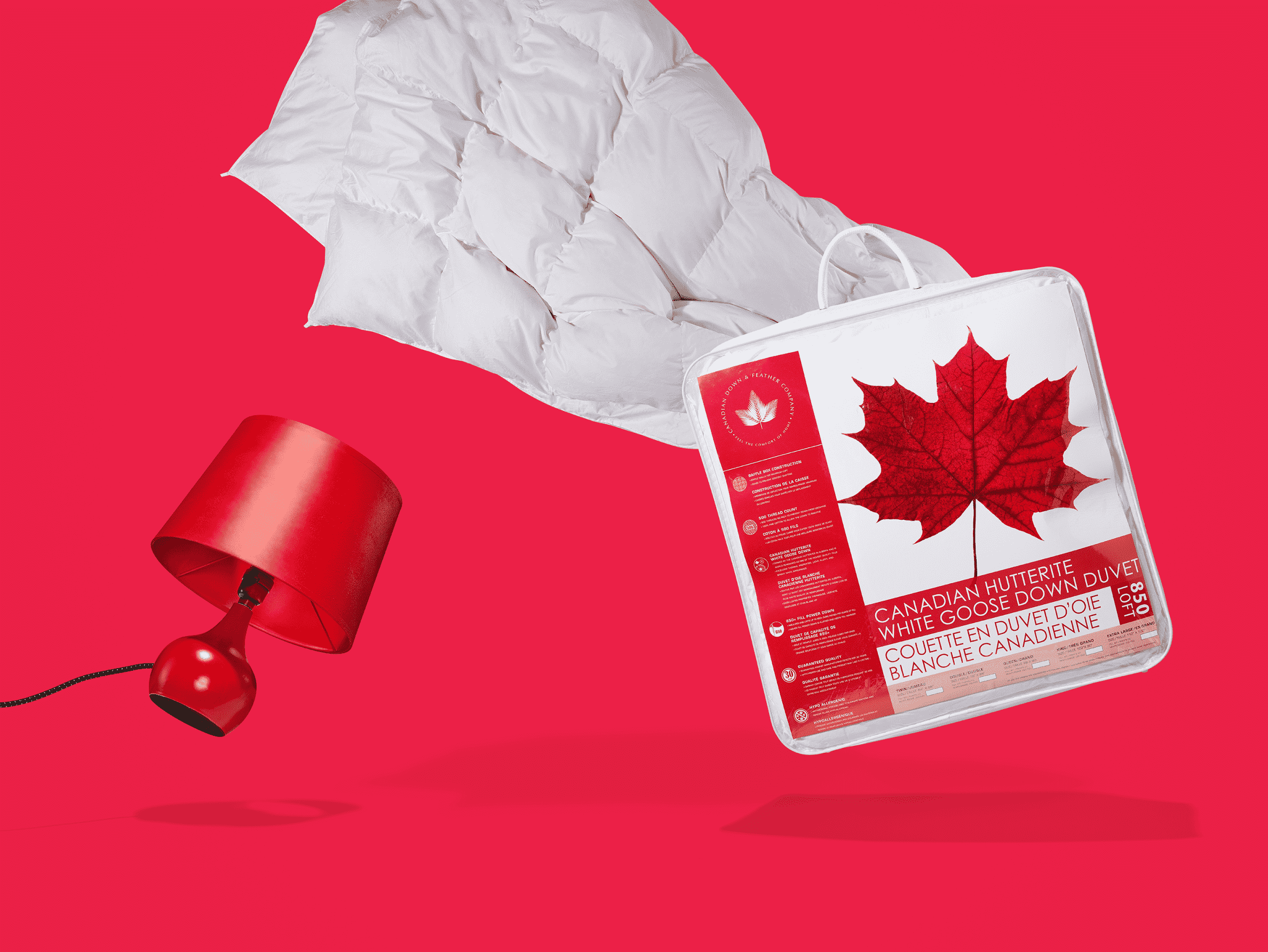 Devant un fond rouge, une couette de duvet d’oie blanche de Canadian Down and Feather emballée, à côté d’une lampe de chevet rouge.
