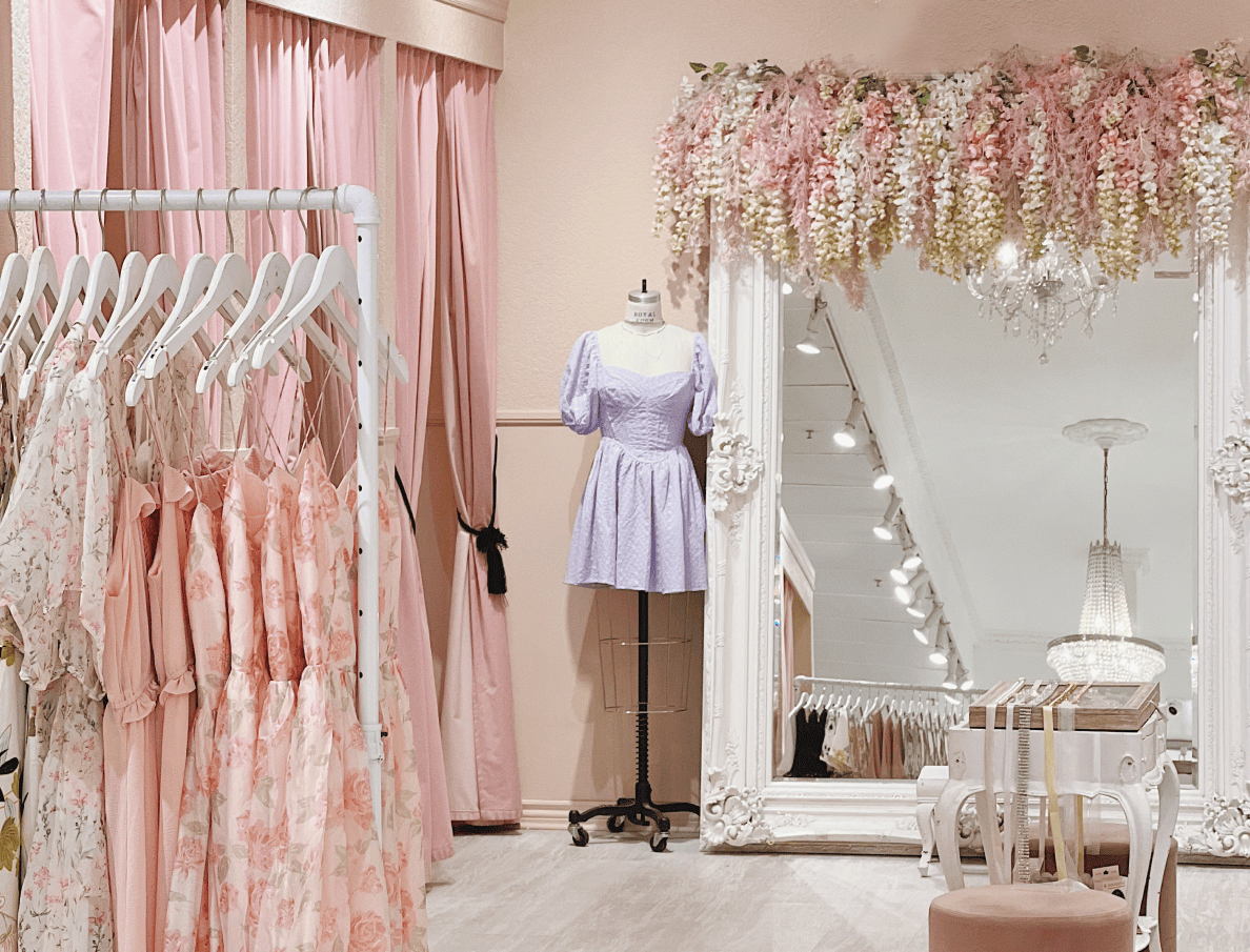 L’extérieur des salles d’essayage d’une Boutique 1861 garnies de rideaux roses et doté d’un superbe miroir de plain-pied. À l’avant plain, de robes roses fleuries sur un présentoir à vêtements. 