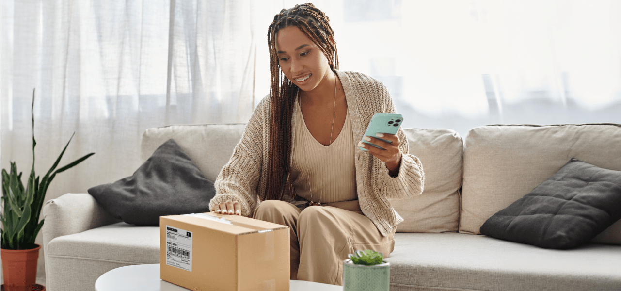 A smiling woman looks at her smart phone as she holds a package.