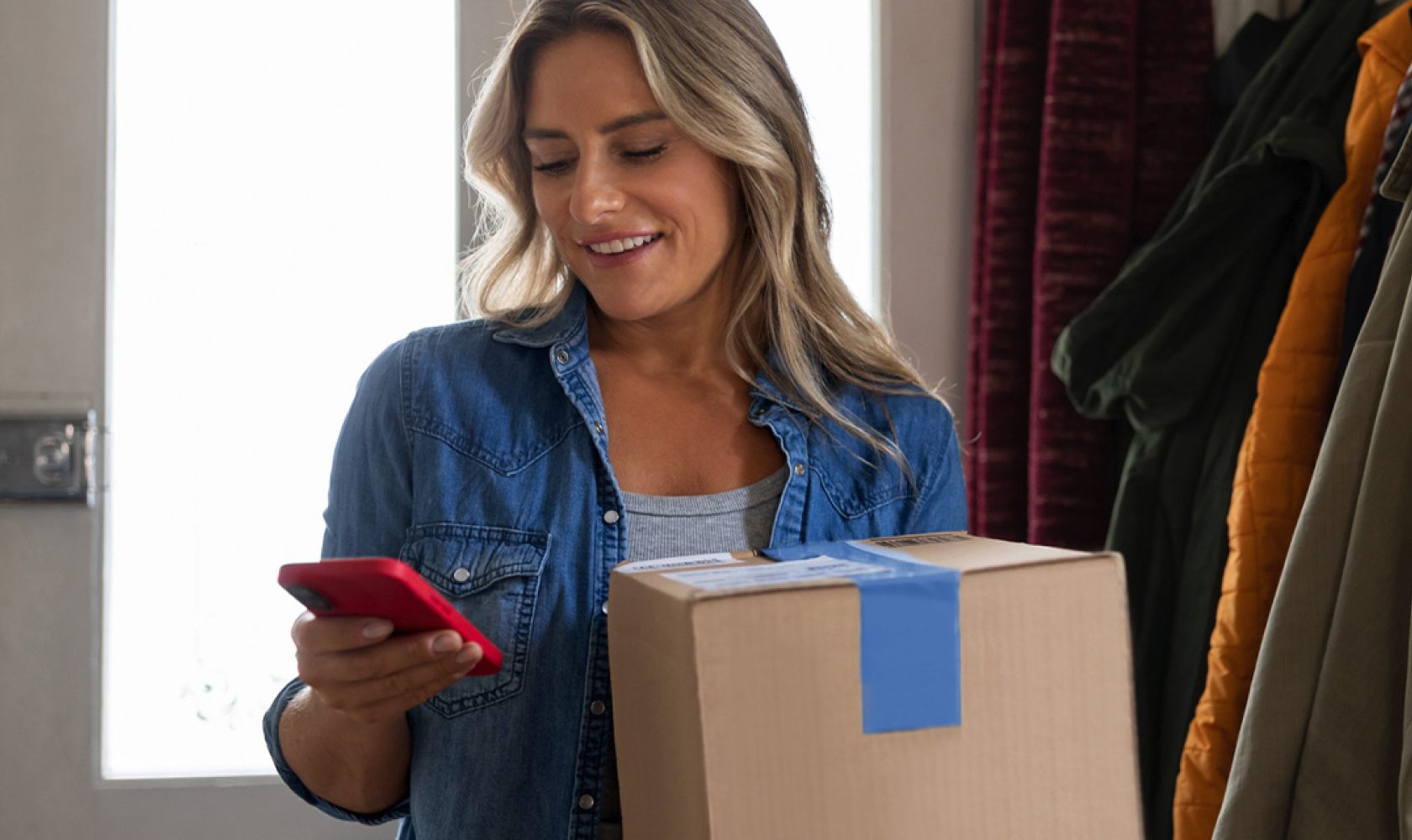 A smiling woman looks at her smart phone as she holds a package.