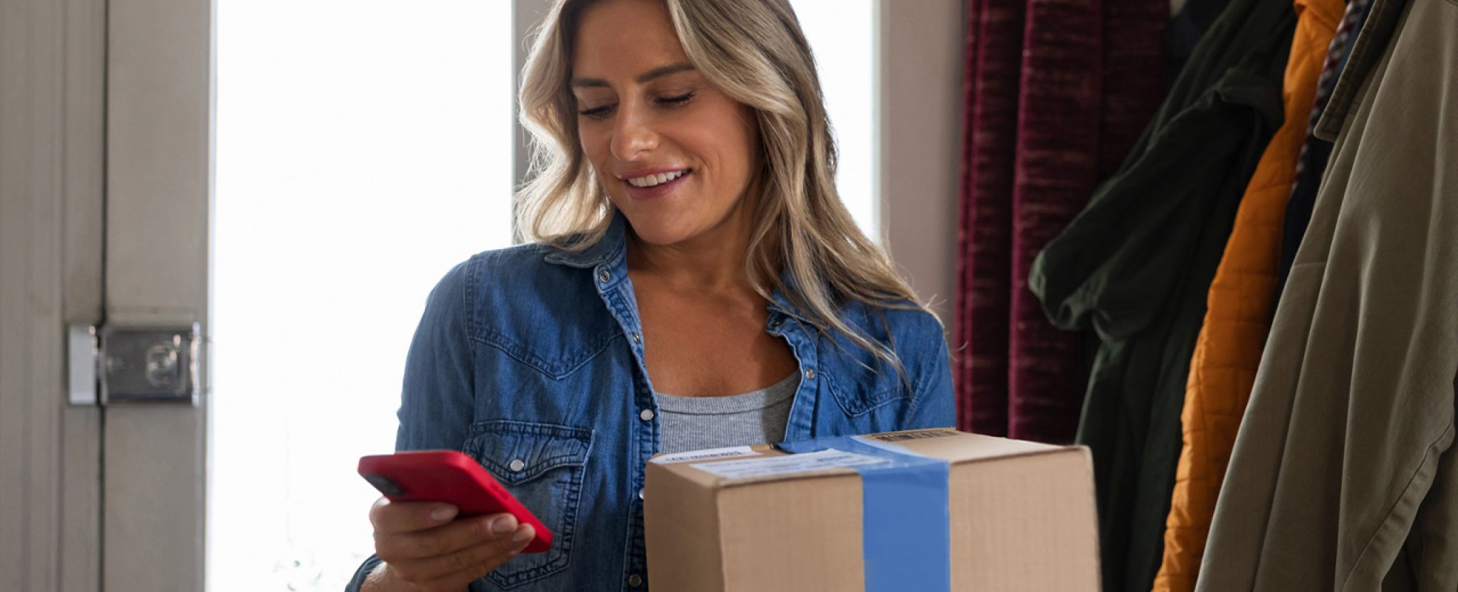 A smiling woman looks at her smart phone as she holds a package.