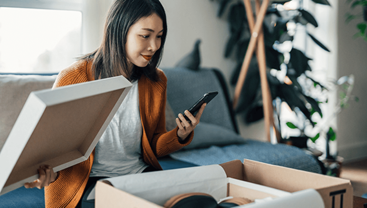 A woman looks at her smartphone at home while she opens a package.