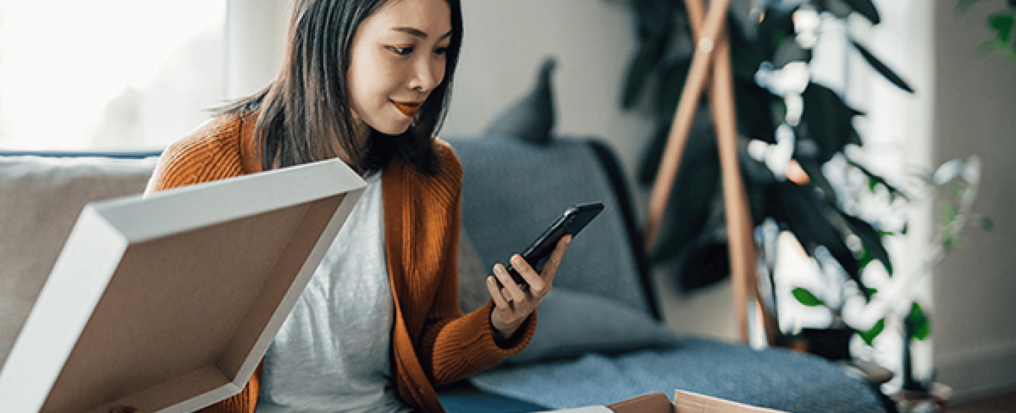 A woman looks at her smartphone at home while she opens a package.