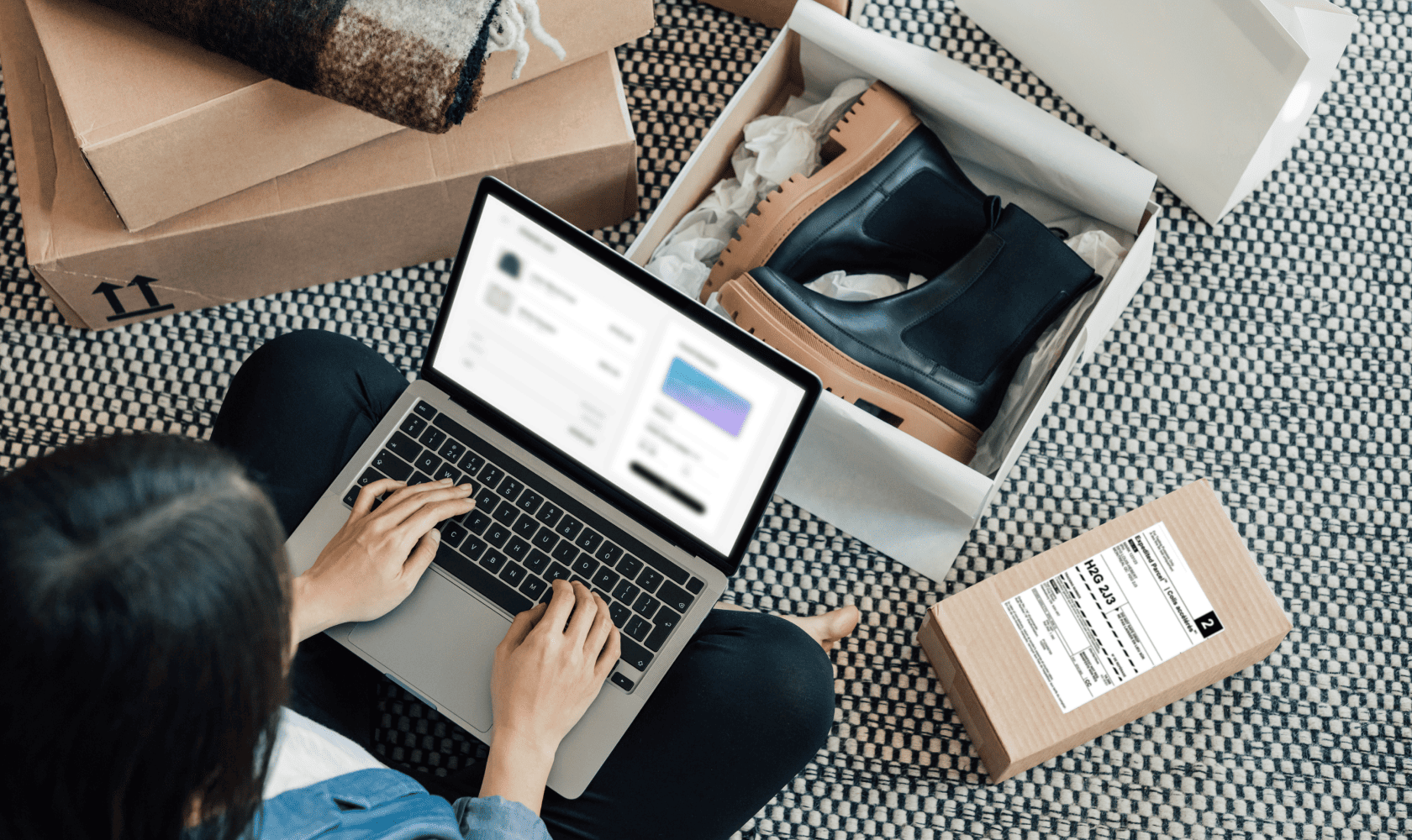 A woman sits on the floor with a laptop, preparing a pair of boots for an online return.