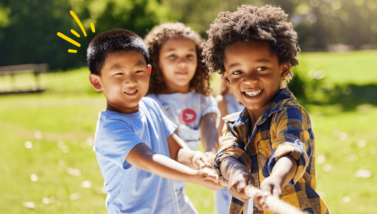 Children playing tug-of-war outdoors, pulling a rope together on a sunny day with grass and trees in the background.