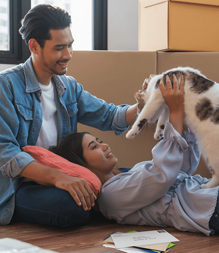 A young couple play with a cat on the floor of their home. Moving boxes are stacked behind them. A young couple play with a cat on the floor of their home. Moving boxes are stacked behind them.