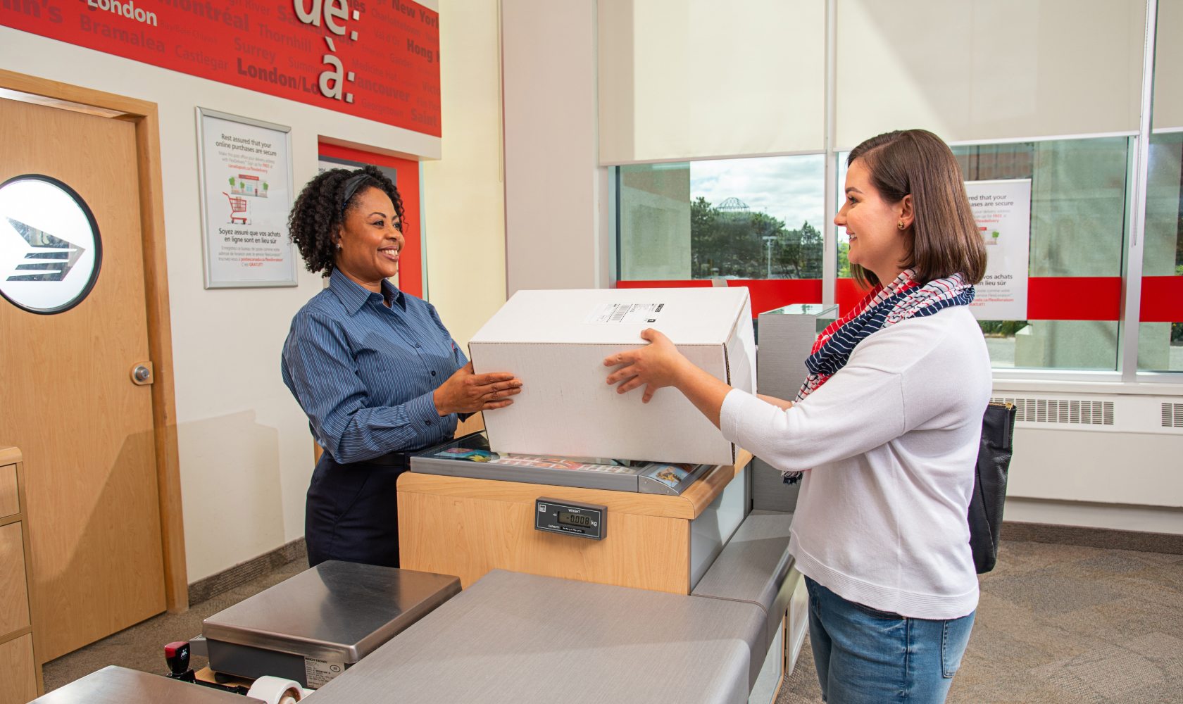 A smiling Canada Post employee hands a package to a smiling customer at a post office.
