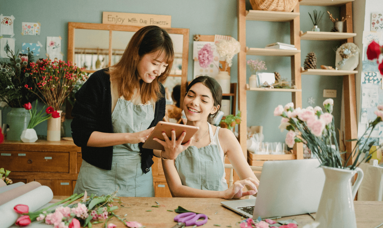 2 women smile as they look at the same tablet computer in a flower shop. They both wear aprons.