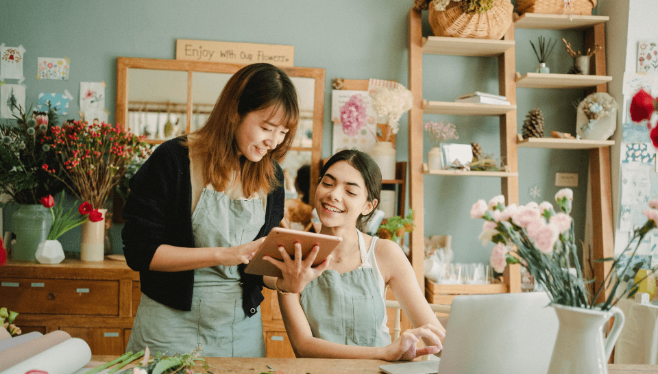 2 women smile as they look at the same tablet computer in a flower shop. They both wear aprons.