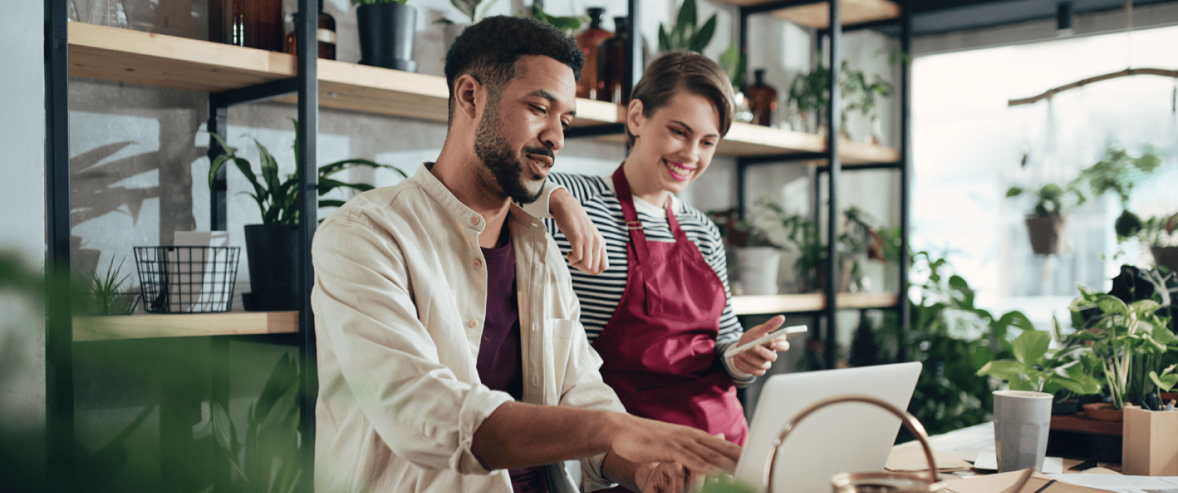 A woman in an apron leans on the shoulder of a man who points at a laptop screen in a plant shop.