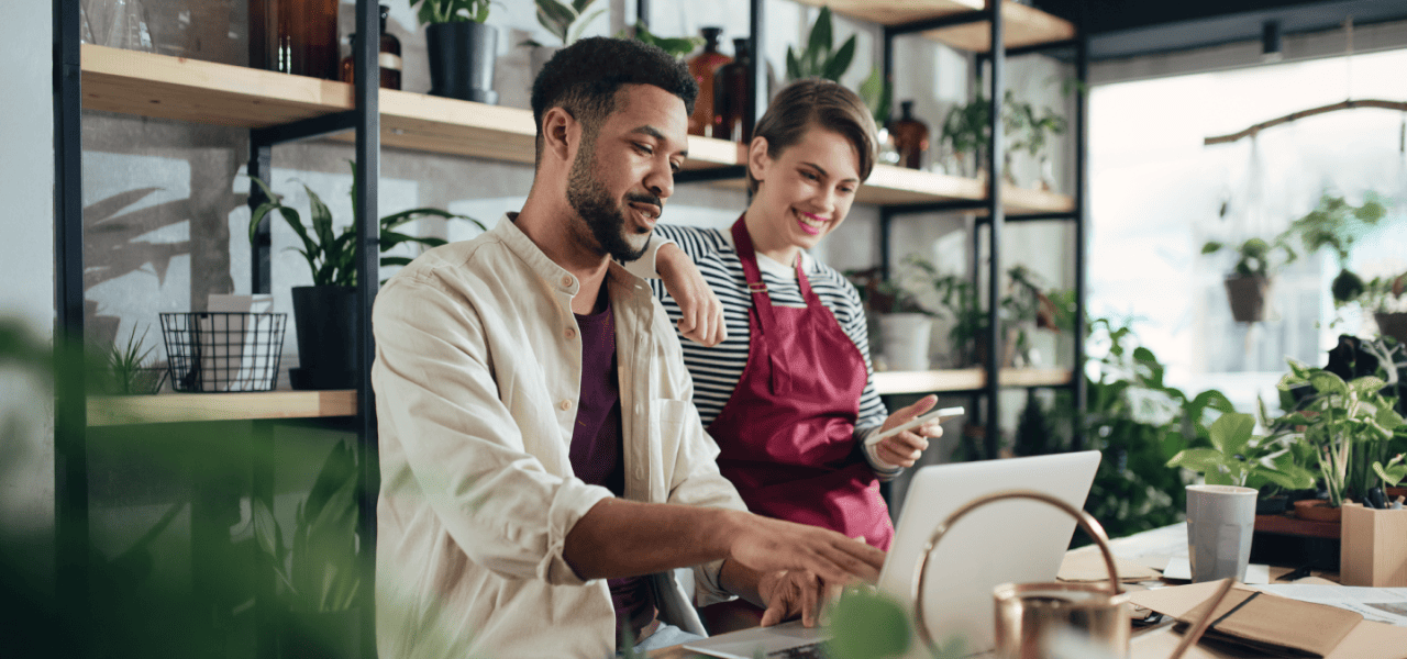 A woman in an apron leans on the shoulder of a man who points at a laptop screen in a plant shop.