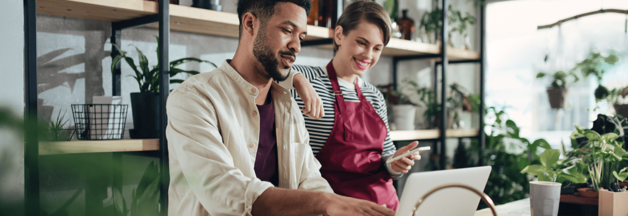 A woman in an apron leans on the shoulder of a man who points at a laptop screen in a plant shop.