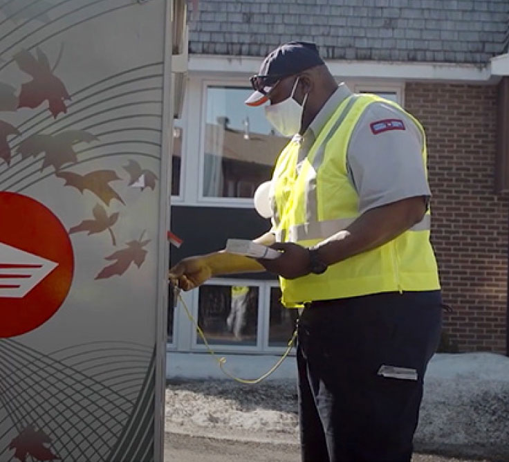 A Canada Post employee wears a yellow safety vest and a face mask. He unlocks a Canada Post delivery vehicle.