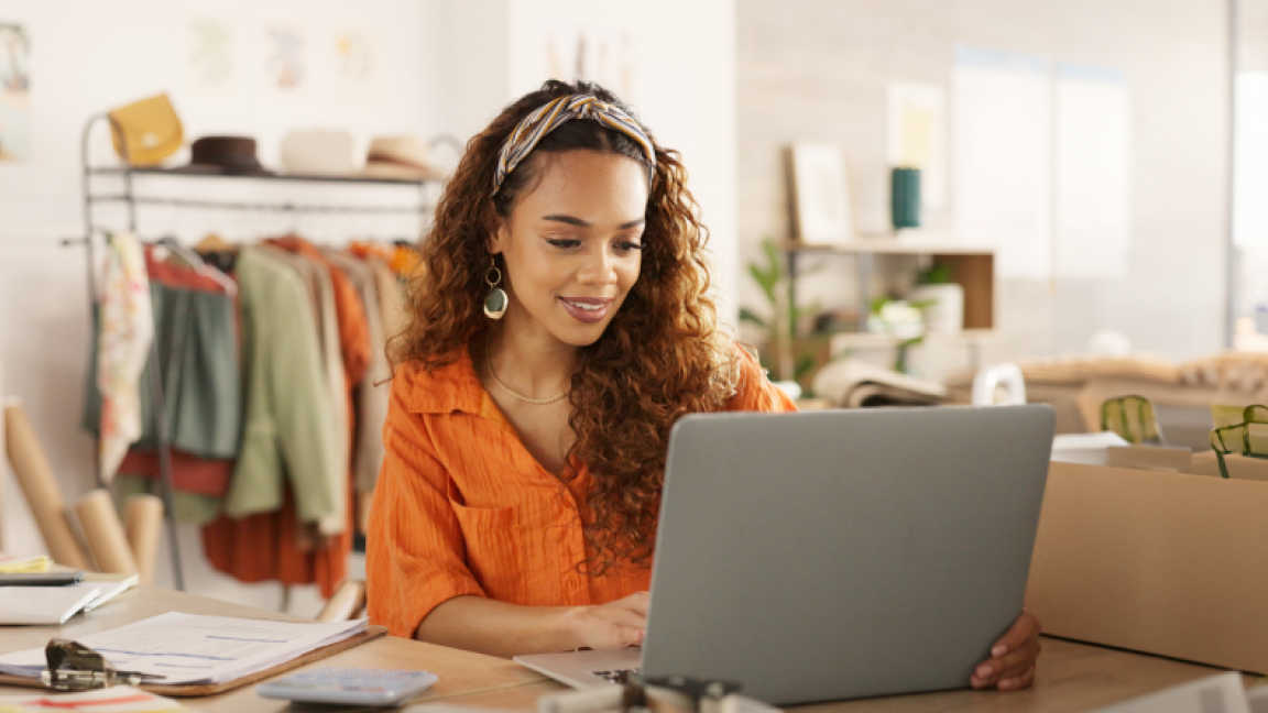 A woman sits at a desk working on a laptop with a clothing rack and shipping supplies in the background.  