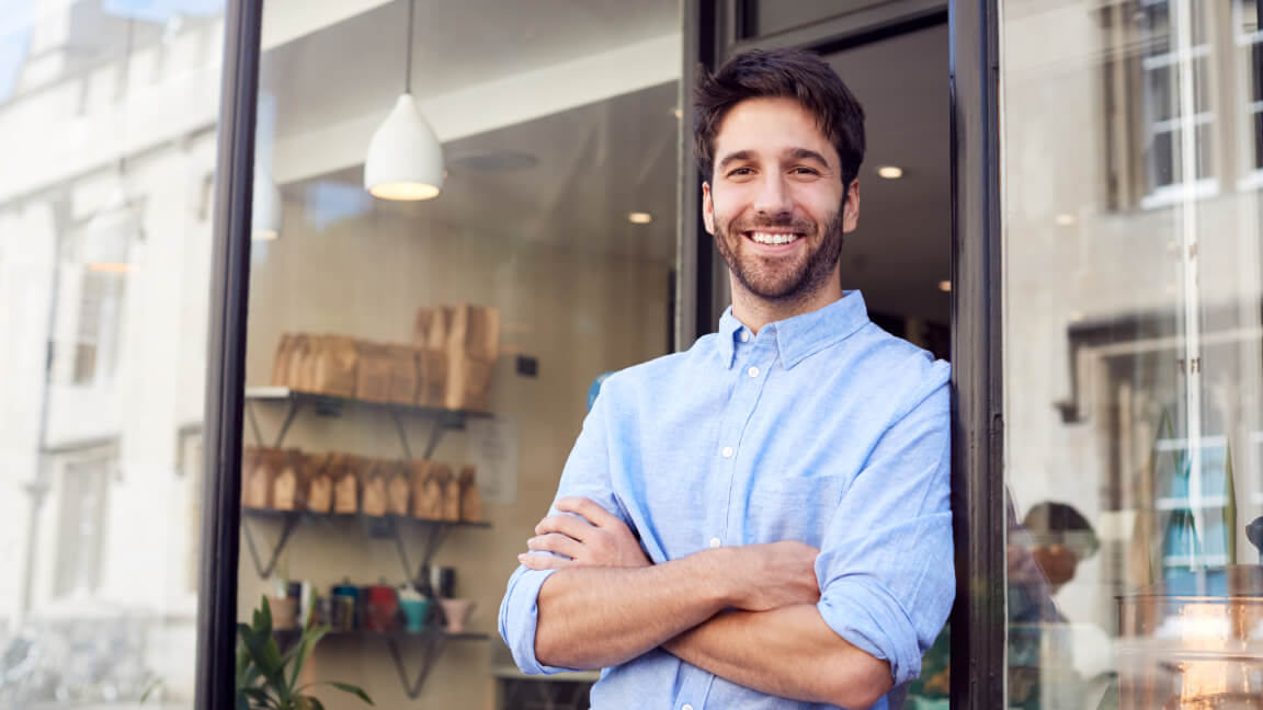 A smiling man has his arms crossed as he casually leans against the open door of a small shop. 