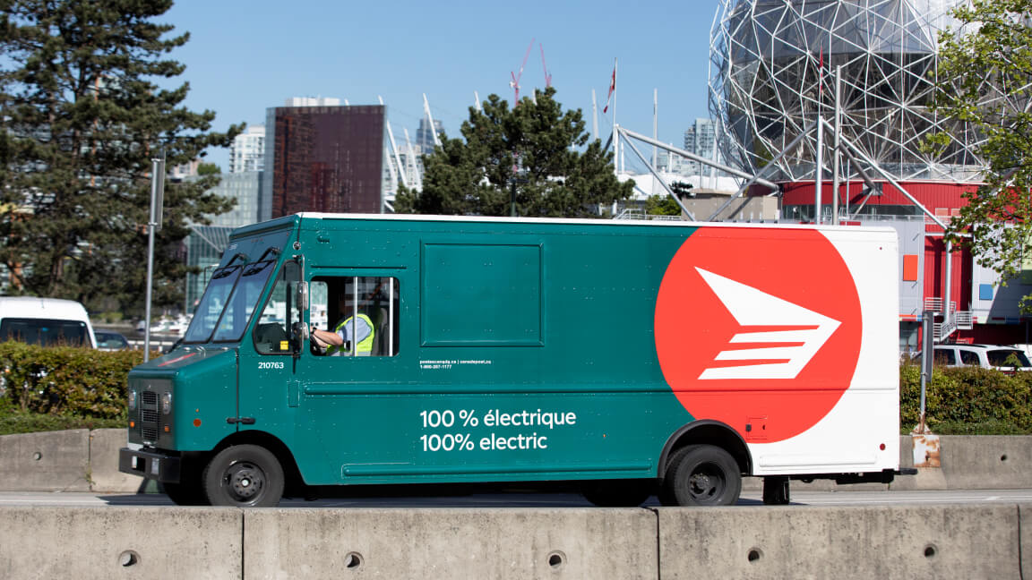 A 100% electric Canada Post truck drives along a road with the Vancouver, B.C. skyline in the background