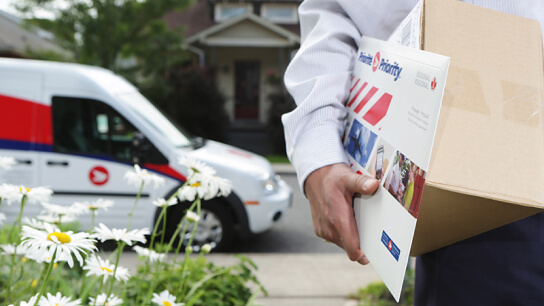 A letter carrier holds a shipping box and a bubble mailer. Their Canada Post vehicle is parked nearby. A letter carrier holds a shipping box and a bubble mailer. Their Canada Post vehicle is parked nearby.