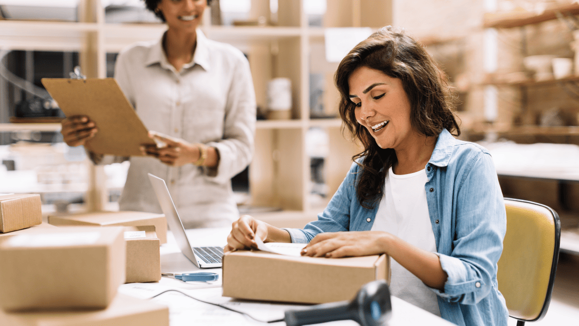 A woman sits at a desk and affixes a shipping label to a box. A woman stands near her holding a clipboard.