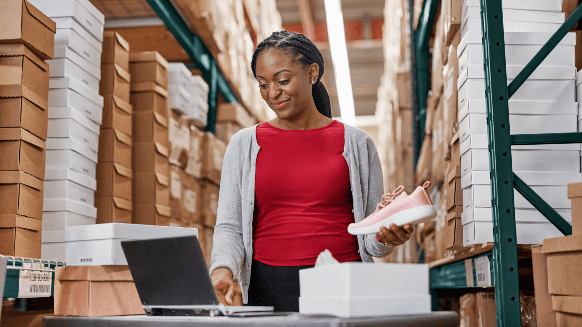A woman types on a laptop as she stands in a large warehouse full of boxes stacked high.