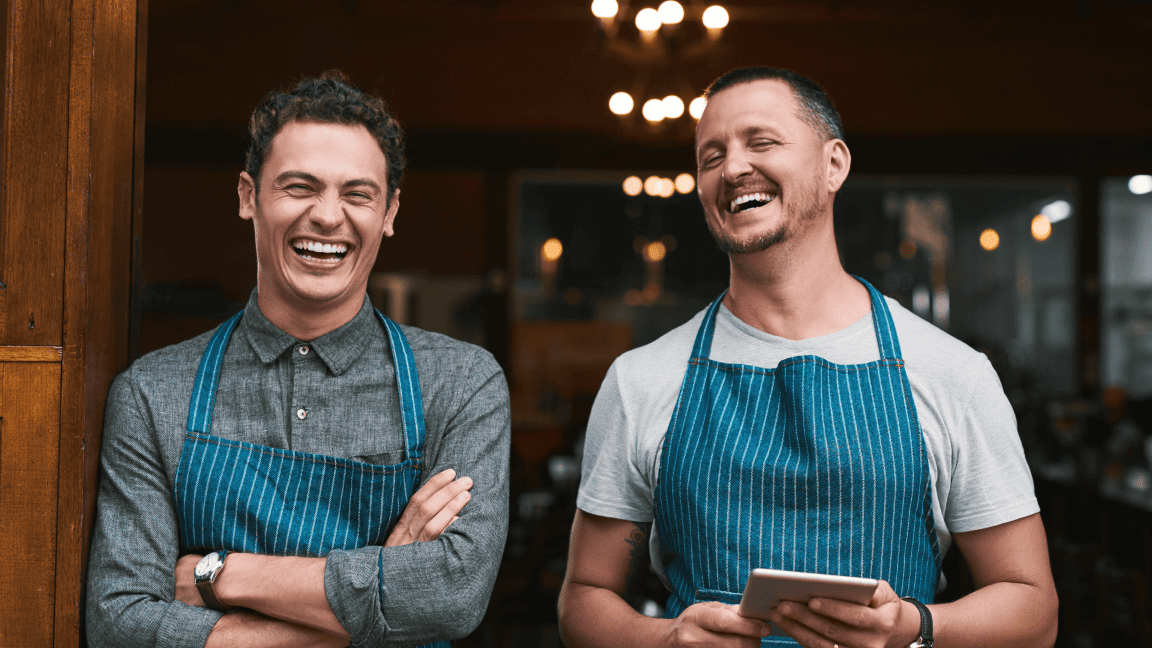 Two men in aprons smile and laugh. One holds a tablet.