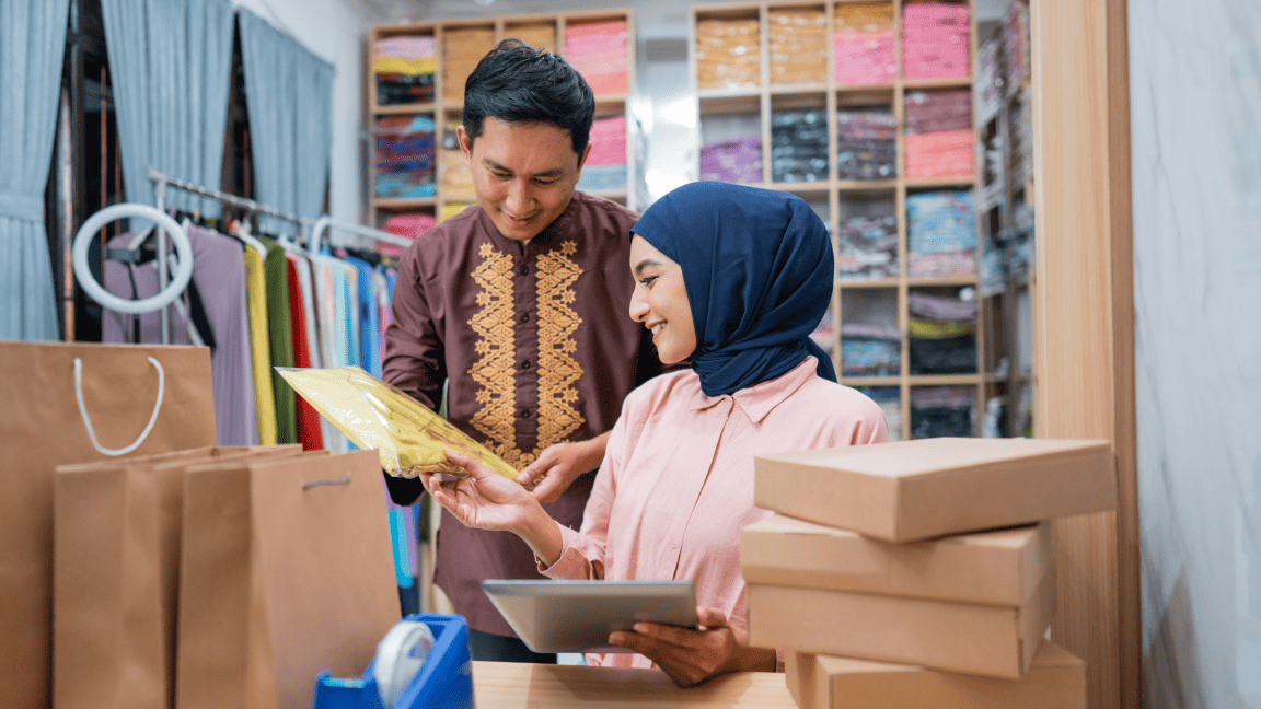 A woman wearing a head covering sits in a clothing store room full of boxes and accepts an envelope from a man who stands behind her.