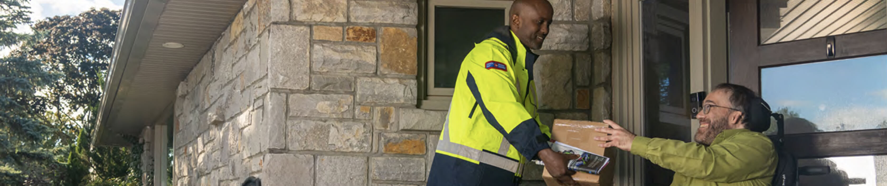 Un agent de livraison de Postes Canada tend un colis à un homme en fauteuil roulant devant sa porte d’entrée. 