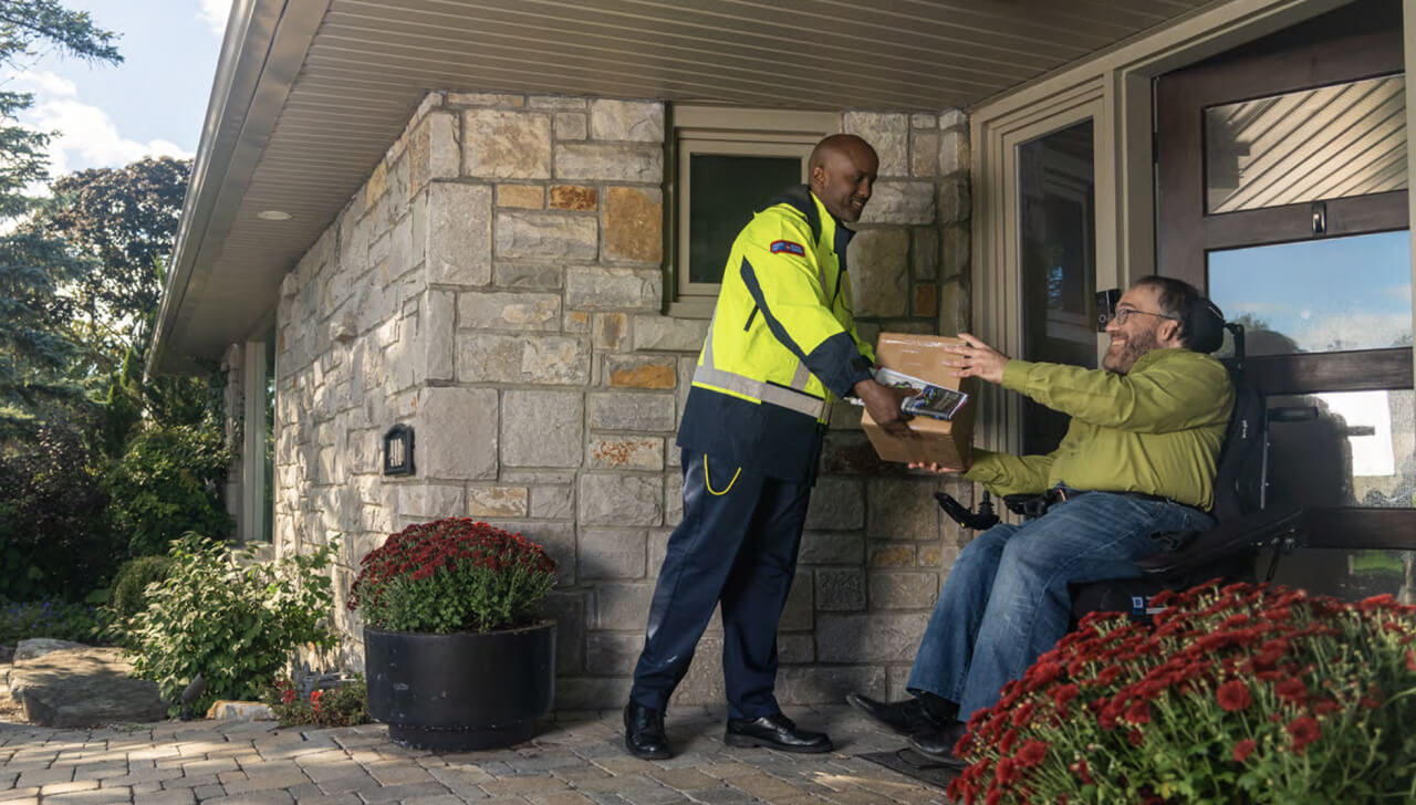 Un agent de livraison de Postes Canada tend un colis à un homme en fauteuil roulant devant sa porte d’entrée. 