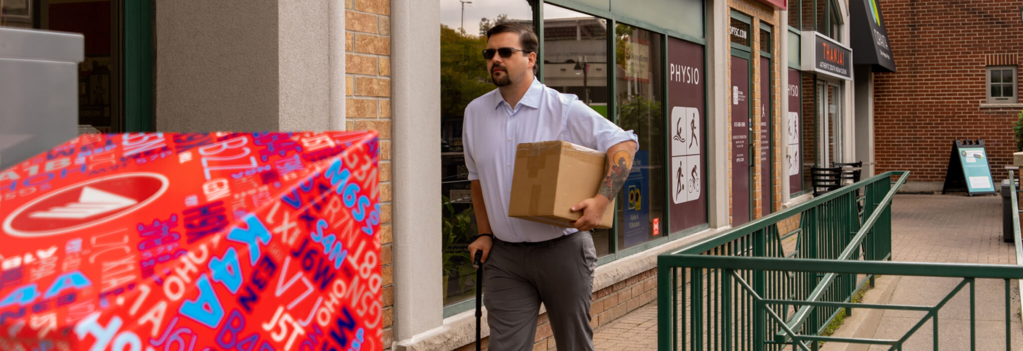 Un homme transportant un colis marche avec une canne sur une rampe de trottoir le long d’un magasin.