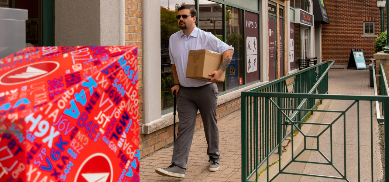 Un homme transportant un colis marche avec une canne sur une rampe de trottoir le long d’un magasin.