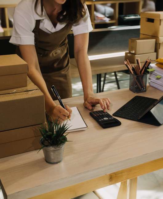 Une propriétaire d’une petite entreprise, vêtue d’un tablier, travaille debout derrière une table. Elle utilise un ordinateur et une calculatrice. Il y a des colis à côté d’elle.