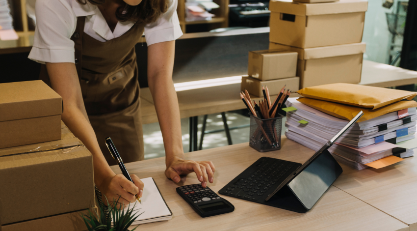 Une propriétaire d’une petite entreprise, vêtue d’un tablier, travaille debout derrière une table. Elle utilise un ordinateur et une calculatrice. Il y a des colis à côté d’elle.