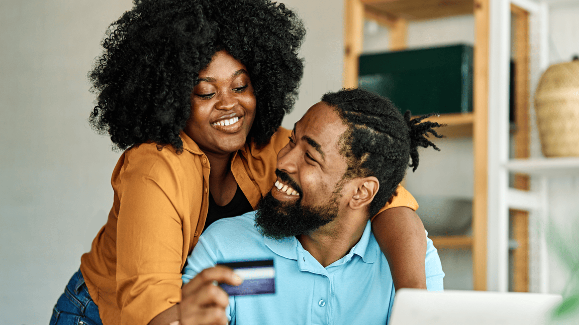 Un couple aimant sourit. L’homme est assis devant un ordinateur portable et tient une carte. La femme se tient derrière lui et l’enlace.